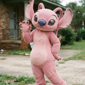 Peut inclure: Un costume en peluche rose représentant le personnage Angel de Lilo et Stitch. La tenue présente de grandes oreilles, un nez bleu, des yeux sombres et une tache rose plus claire sur la poitrine. Le personnage fait un signe de la main.