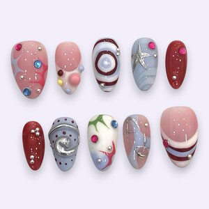 Peut inclure: Un ensemble de dix faux ongles présentant des designs variés avec des bases roses, bleues et rouges, agrémentés d'éléments 3D tels que des spirales métalliques argentées, des étoiles, des petites sphères et des strass colorés incrustés.