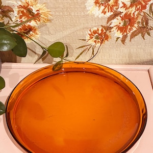 May include: A round, amber-coloured glass tray with a smooth, translucent surface and a slightly raised rim. The tray features a deep, warm orange hue and a polished finish, reflecting light across its circular form and flat base.