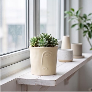 May include: A cream-colored ceramic planter with a minimalist embossed face design on the front, containing a cluster of green succulent plants. The cylindrical pot features a smooth matte finish and is positioned upright on a flat surface.