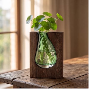 May include: A clear, teardrop-shaped glass vase is set within a rectangular dark brown wooden frame. The vase contains water and a green plant with round, flat leaves. The wooden frame has a visible grain and a smooth, flat-topped design.