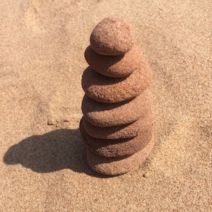 Puede incluir: Una pila de seis piedras lisas de color marrón rojizo, equilibradas unas sobre otras sobre una superficie arenosa.