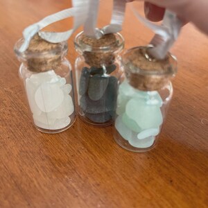 May include: Three small clear glass bottles with cork stoppers, each filled with smooth, rounded stones in white, dark grey, and pale green. Each bottle features a white ribbon loop attached to the top for hanging, creating a set of ornaments.