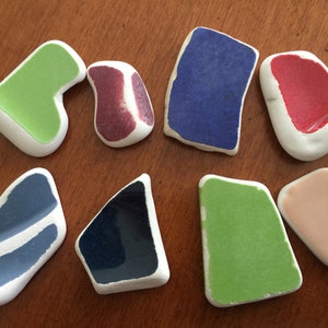 以下が含まれることがあります： 緑、青、赤、ピンク、紫など、さまざまな形と色のセラミックタイル8枚。タイルは木製の表面に並べてあります。