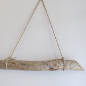 May include: A natural wood branch with a rough, textured surface is suspended from a white wall by a length of twine. The branch is approximately 12 inches long.