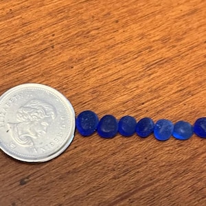May include: A silver coin next to a line of small, oval, blue glass beads. The beads are arranged in a row on a wooden surface. The coin has a profile of a person on it.