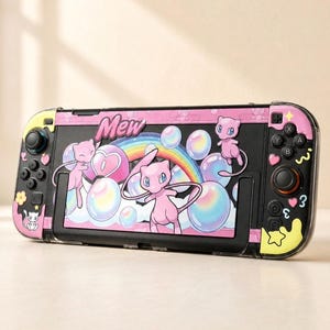 Può includere: Una console di gioco portatile con una skin personalizzata raffigurante il personaggio rosa Mew, un arcobaleno e bolle iridescenti. Il design include la scritta Mew, piccoli cuori, stelle e fiori su una base nera con pulsanti e joystick neri.