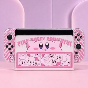 以下が含まれることがあります： ピンクと白のストライプ柄にカービィのキャラクターが描かれたゲーム機用ドックとコントローラーのセット。ドックには「PINK PUFFY POWERFUL」の文字と、複数のカービィが描かれたグラフィックが表示されています。コントローラーにもカービィのアイコンがあしらわれています。