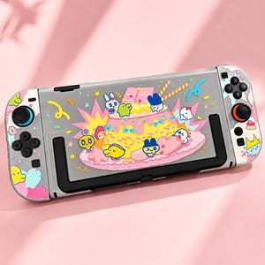 May include: A handheld gaming console featuring a clear protective case decorated with colorful cartoon characters, a large yellow pudding dessert, pink frosting, and confetti patterns. The device includes black joysticks and buttons.