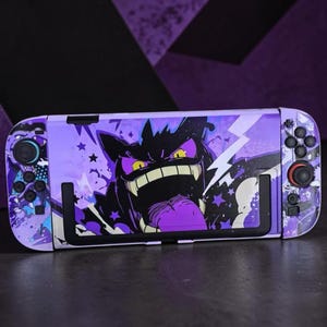 Könnte beinhalten: Eine tragbare Spielkonsole mit einer violett-schwarzen Hülle, die eine grafische Illustration einer Kreatur mit weit geöffnetem Mund, gelben Augen und einer langen Zunge zeigt. Das Design umfasst Sternmuster und Blitze auf der Vorderseite.