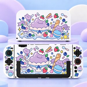 May include: A white gaming console dock and handheld controller set featuring a colourful pattern of purple blob characters, blue macarons, strawberries, blueberries, and whipped cream desserts with wafer sticks on a white background.