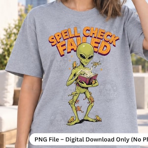 May include: A heather gray t-shirt featuring a graphic of a green alien reading a book with the text "SPELL CHECK FAILED" in orange letters above the alien. Several small frogs are positioned around the alien. The design includes a logo in the top right corner.