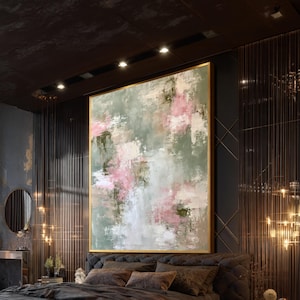 Puede incluir: Un gran cuadro cuadrado sobre lienzo que presenta una composición abstracta de pinceladas mezcladas en verde salvia, rosa suave y blanco con textura visible, encerrado en un marco dorado fino y montado en una pared oscura sobre un cabecero capitoné.