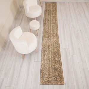 May include: A long, narrow rectangular rug featuring a faded, distressed pattern in shades of beige, brown, and muted gray. The rug displays an intricate, repeating floral and geometric motif throughout its surface, with a distinct border design.