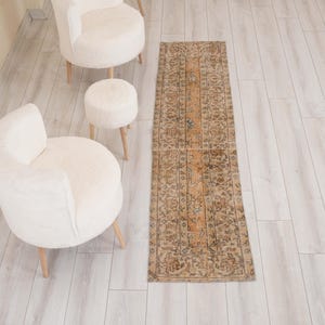 Könnte beinhalten: Ein langer, rechteckiger Läufer mit einem verblassten, komplizierten Blumenmuster in Beige-, Braun- und gedämpften Orangetönen, platziert neben zwei cremefarbenen, abgerundeten Polstersesseln und einem kleinen passenden runden Hocker.