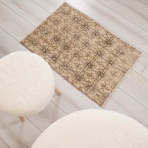 Peut inclure: Un tapis rectangulaire au fond beige avec un motif géométrique répétitif brun foncé, disposé à côté de trois tabourets ronds, blancs, duveteux et rembourrés avec des pieds en bois clair sur un sol en bois de couleur claire.