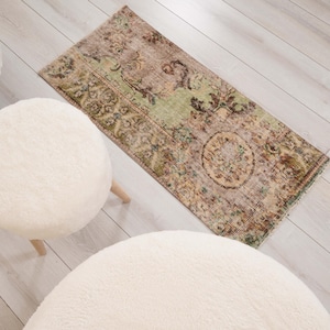 May include: A rectangular area rug featuring a faded, distressed pattern in shades of sage green, brown, and beige, with intricate floral and geometric motifs. The rug is positioned near two round, white, fuzzy, upholstered stools with wooden legs.