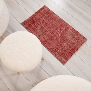 Puede incluir: Una alfombra rectangular con un patrón rojo y blanco desgastado y textura envejecida, colocada junto a tres taburetes redondos, blancos, esponjosos y tapizados con patas de madera clara sobre un suelo con aspecto de madera clara.