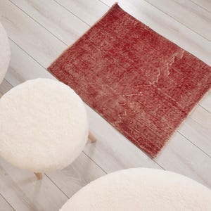 May include: A rectangular rug featuring a deep red color with a distressed, weathered texture and a subtle white zigzag pattern running across the surface. The rug is positioned near three round, white, fuzzy, textured stools.
