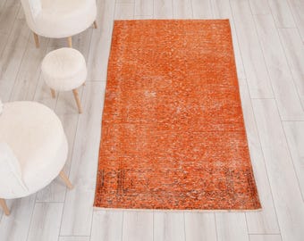 Puede incluir: Una alfombra rectangular que presenta un campo de color naranja vibrante con una textura desgastada y descolorida, junto con intrincados patrones de borde en gris carbón oscuro. La alfombra muestra un diseño tejido tradicional con signos visibles de uso.