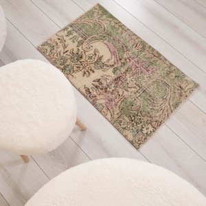 Puede incluir: Una alfombra rectangular con un patrón floral descolorido en tonos verde salvia, crema y rosa apagado, situada junto a tres taburetes redondos, blancos y esponjosos con patas de madera clara sobre un suelo de aspecto madera.