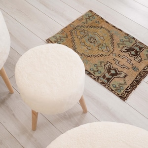 Könnte beinhalten: Drei runde Hocker aus weißem Kunstfell mit hellen Holzbeinen, angeordnet um einen kleinen rechteckigen Teppich mit geometrischem Muster in Beige-, Braun- und Petroltönen mit dunkelbraunen Rändern und gefransten Kanten.