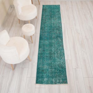 May include: A long, rectangular runner rug in a deep teal color with a faded, distressed pattern. The rug features intricate, worn-in details throughout its surface, showcasing a vintage-inspired aesthetic with varied shades of blue and green.