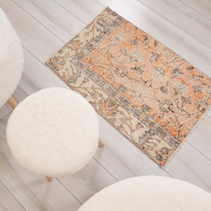 Könnte beinhalten: Ein rechteckiger Teppich mit einem verblassten Blumenmuster in Orange, Creme und Schieferblau sowie einer dekorativen Bordüre, begleitet von drei runden, weißen, strukturierten und flauschigen Hockern mit hellen Holzbeinen um den Teppich.
