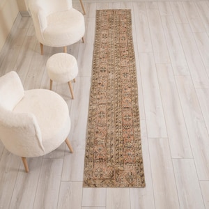 May include: A long, narrow runner rug featuring a faded, intricate pattern in muted shades of pink, brown, and beige. The rug is rectangular with a repeating geometric and floral design, positioned on a light-colored wood floor near two chairs.