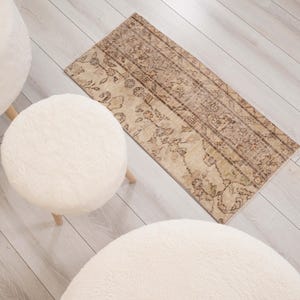 May include: A rectangular rug featuring a faded, intricate brown and beige floral pattern, positioned alongside three round, cream-colored stools with textured, fuzzy upholstery and light-toned wooden legs arranged on a light-colored floor.