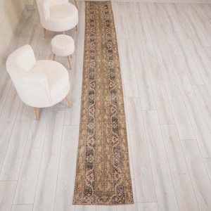 May include: A long, narrow rectangular rug featuring a distressed, faded pattern in shades of brown, tan, and charcoal gray. The rug displays a repeating geometric and floral motif throughout its length, with a dark border framing the design.