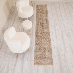 May include: A long, narrow runner rug featuring a faded, intricate pattern in shades of beige, brown, and muted grey. The rug is rectangular with a flat-weave texture and displays a repeating floral and geometric design across its entire surface.