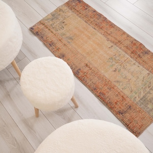Può includere: Un tappeto rettangolare con un motivo sbiadito e usurato nei toni dell'arancione, beige e blu tenue, posizionato accanto a tre sgabelli rotondi imbottiti color crema con gambe in legno chiaro su un pavimento di colore chiaro.