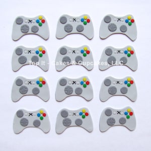 May include: Twelve gray and white video game controller cake toppers with colorful buttons. Top It - Cakes & Cupcakes, LLC.
