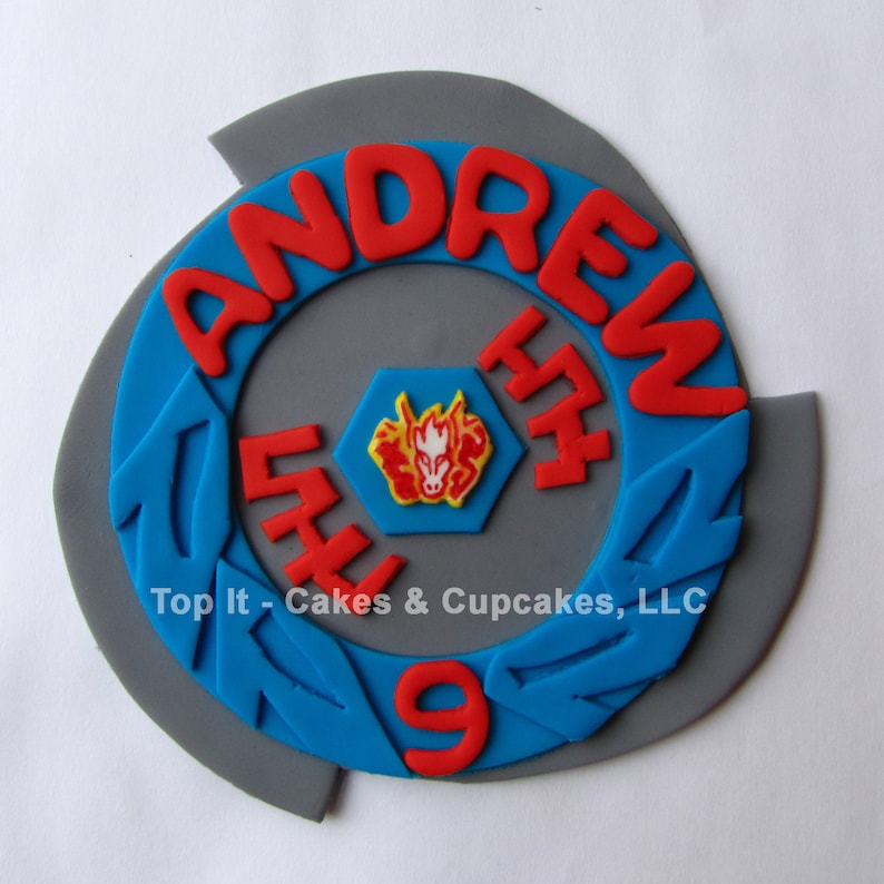 May include: A gray and blue cake topper shaped like a Beyblade with the name "Andrew" and the number "9" in red frosting. The center of the Beyblade has a red, yellow, and white design.