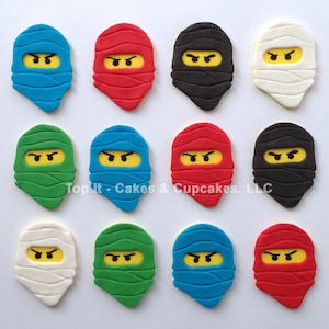 May include: Twelve fondant cake toppers shaped like ninja masks. The masks are in various colors, including blue, red, green, black, and white. Each mask has a yellow face with black eyes and eyebrows.