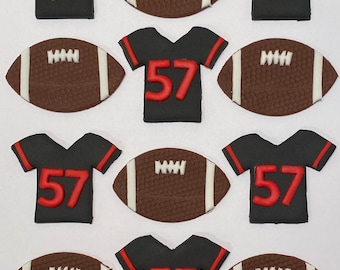 May include: Twelve edible cake toppers featuring football jerseys and footballs. The jerseys are black with red trim and the number 57. The footballs are brown with white laces.