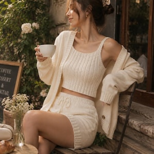 May include: A cream-colored three-piece knit set consisting of a ribbed crop top with spaghetti straps, high-waisted shorts with a drawstring waist, and a long-sleeved open cardigan with buttons. The fabric features a consistent vertical knit texture.