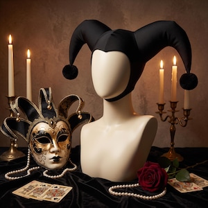Könnte beinhalten: Eine schwarze Narrenkappe mit zwei langen, geschwungenen Spitzen, die in schwarzen Bommeln enden, präsentiert auf einem Schaufensterpuppenkopf. Daneben liegt eine dekorative venezianische Maske mit Gold- und Schwarzverzierungen, Perlen und Spielkarten.