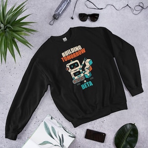 May include: A black long-sleeve sweatshirt featuring a graphic of a small robot stacking blue and orange cubes. The text above the robot reads "BUILDING TOMORROW" in orange, and the text below reads "IN BETA" in light blue, with a code symbol on the chest.