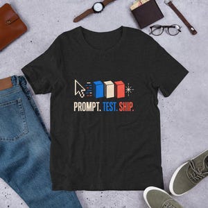 May include: A black short-sleeve t-shirt featuring a graphic design with a white pixelated cursor, three colored blocks in blue, white, and red, and the text PROMPT. TEST. SHIP. printed in white, blue, and red lettering across the chest area.