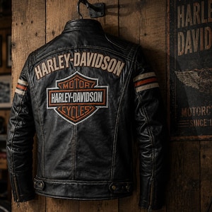 May include: A black leather jacket featuring the Harley-Davidson logo on the back. The design includes a large embroidered shield emblem with the text Harley-Davidson Motor Cycles, plus orange and white stripes on the sleeves and a collar.