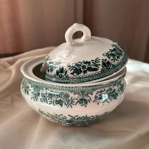 Pode incluir: Um açucareiro de cerâmica branca com tampa, apresentando um padrão floral verde-escuro ao redor da borda e do corpo. A tampa possui uma pequena alça em forma de laço no topo e a tigela tem um formato arredondado com uma borda festonada.