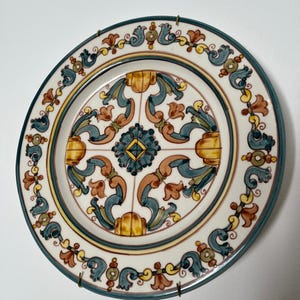 Pode incluir: Um prato decorativo redondo de cerâmica com um padrão simétrico pintado à mão em tons de azul, amarelo e terracota sobre um fundo creme. O design inclui motivos florais centrais e elementos de volutas, com uma borda azul escura.