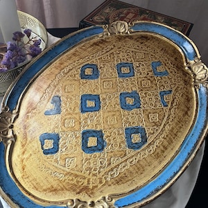 Op de afbeelding: Een ovaal decoratief dienblad met een verweerde gouden afwerking en een ingewikkeld reliëfpatroon. De rand is geschilderd in levendig blauw met gouden accenten en het midden toont een raster van blauwe vierkanten op de textuurrijke gouden ondergrond.