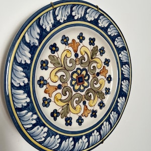 Pode incluir: Um prato decorativo redondo de cerâmica com um motivo floral central de volutas amarelas, verdes e azuis. A borda é pintada com um padrão repetitivo de folhas azuis e brancas, acentuado por linhas amarelas finas, e preso por clipes metálicos.