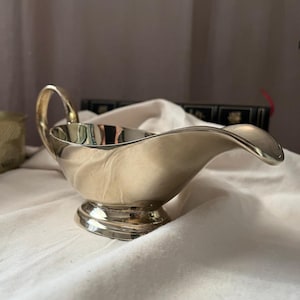 Può includere: Una salsiera color argento lucido con manico curvo, ampio beccuccio e base circolare a piedistallo. La superficie metallica presenta una finitura riflettente con contorni lisci e arrotondati dal design classico e simmetrico.