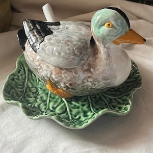 Pode incluir: Um recipiente de cerâmica em forma de pato com tampa, sobre um pires verde em forma de folha. O pato apresenta peito branco, asas cinzentas e pretas, um olho amarelo e um bico laranja. Uma pequena colher de cerâmica branca sai das costas.