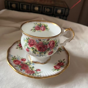 Könnte beinhalten: Ein weißes Porzellan-Tassenset mit Untertasse, verziert mit einem Blumenmuster aus roten Rosen und kleinen blauen Blüten. Die Ränder von Tasse und Untertasse sind goldfarben abgesetzt, und die Tasse besitzt einen verzierten, geschwungenen Henkel.