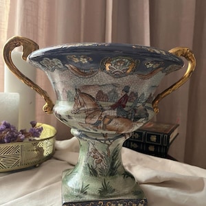 Pode incluir: Um vaso de cerâmica em forma de urna com acabamento craquelado, duas alças douradas ornamentadas e uma cena pintada à mão retratando figuras a cavalo e um cão de caça numa paisagem, com bordas florais azuis e douradas no topo e na base.